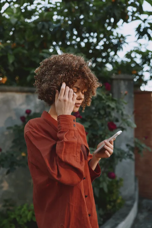 Casual woman in rust sweater checks smartphone outdoors, surrounded by greenery, relaxed pose and thoughtful smile, capturing a warm, lifestyle moment in natural daylight, her curls frame her face