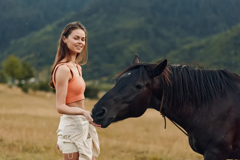 A cheerful young woman feeds a horse in a wide open field, wearing casual summer clothes. She smiles warmly, creating a peaceful and friendly moment in a rural landscape.