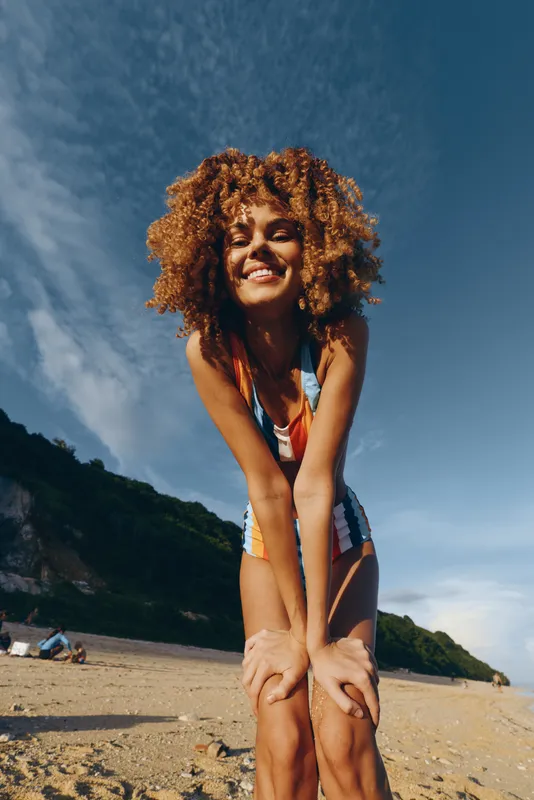 Young woman with curly hair smiles joyfully while bending forward on a sunny beach with a clear blue sky and green hills in the background.