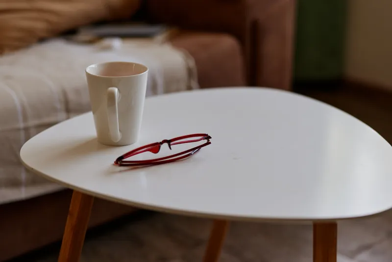 A white round table hosts a mug and a pair of red glasses, with a blurred living room backdrop. A simple home moment depicting quiet morning routine and cozy lifestyle.