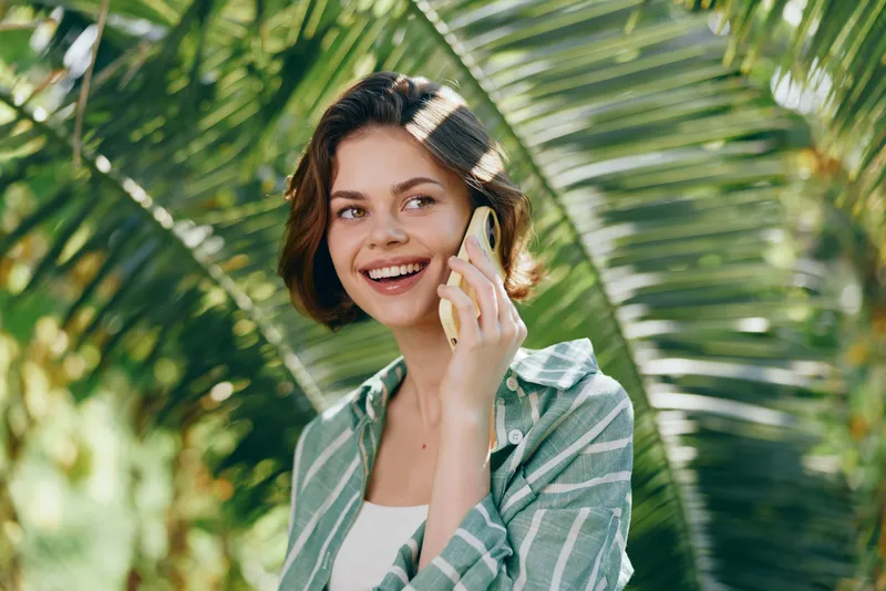 Cheerful woman outdoors chats on a phone amid lush tropical backdrop, wearing a striped green shirt, radiating warmth and positive energy