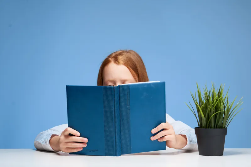 Рыжеволосая девушка сидит за столом с книгой в руках и учит уроки