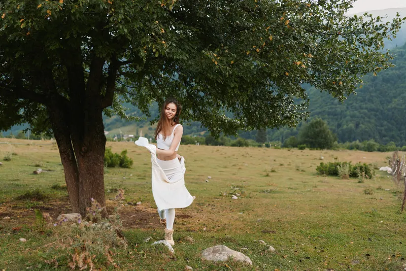 A woman in a white dress spins beside a large tree in an open meadow, capturing a carefree lifestyle moment outdoors, with bright natural light and rural scenery.