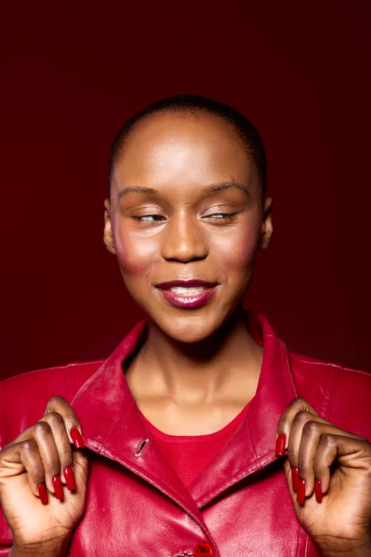 Confident black woman with dark skin wears red leather jacket and matching nails. She looks sideways with a slight smile on dark red background. Concept of fashion, beauty, and style.