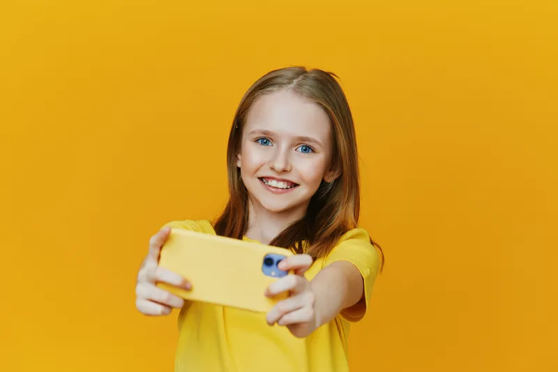 Happy girl taking a selfie with a yellow smartphone against a bright yellow background, wearing a yellow shirt Cheerful emotions and a playful concept
