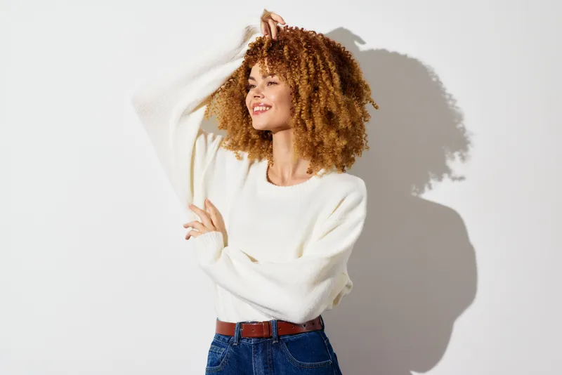 Стильная молодая женщина с вьющимися волосами в мягком белом свитере стоит на светлом фоне