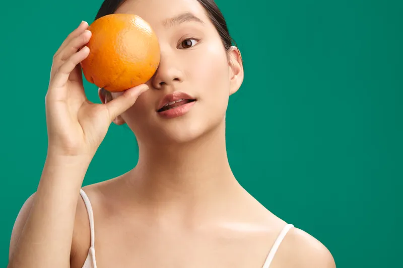 Asian woman holds an orange near her eye in a studio portrait with a vibrant green backdrop, conveying playfulness and freshness in a simple, color driven composition.