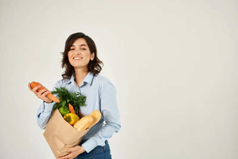 Женщина держит в руках пакет с продуктами, в котором видна морковь и другие овощи, в супермаркете
