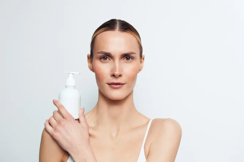Young woman with clear skin holds white cosmetic bottle close to face with gentle expression isolated on light background for skincare or beauty concept.