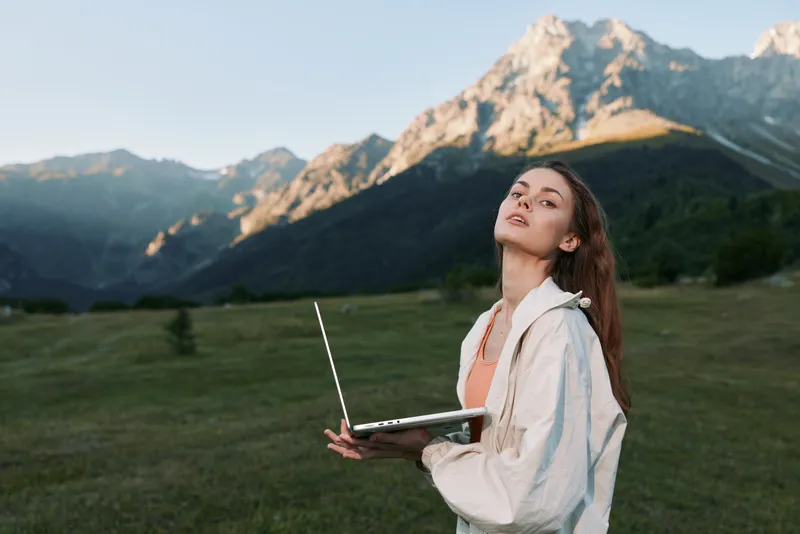 A young woman stands in a vast alpine field with a laptop, gazing upward with a confident, hopeful expression, capturing a moment of remote work in nature.