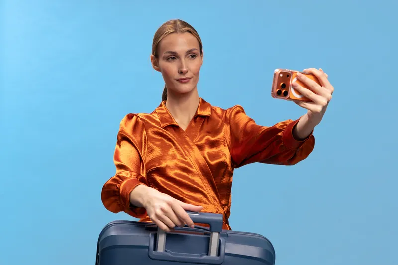 Young woman in orange blouse takes selfie with smartphone while holding suitcase handle on blue background. Concept of travel, vacation, and modern lifestyle with digital communication.