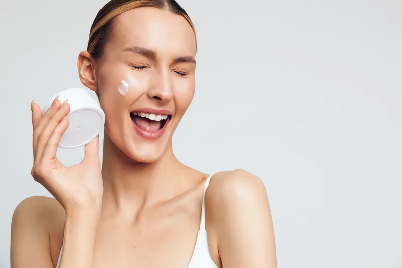 Happy young woman with clean skin applying cream on face holding container smiling with closed eyes isolated on white background skincare beauty concept natural care.