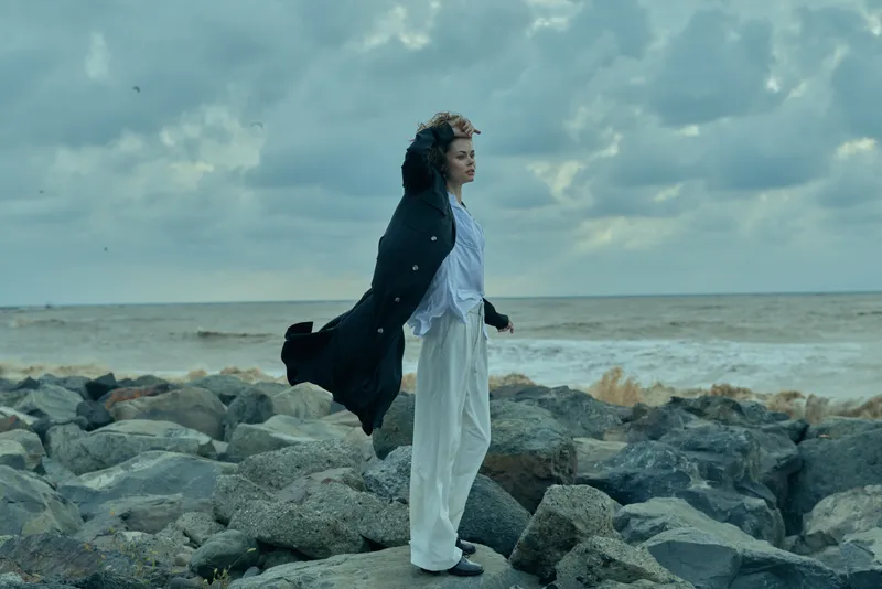 Wind, sea, cloak and dress on rocky shoreline under cloudy sky, flowing fabric captures movement and mood for dramatic coastal fashion and natural scenery