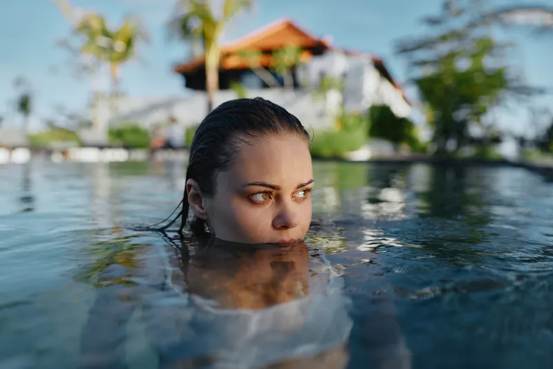 Close up of a pool scene with a woman partially submerged, her face above water, looking toward a tropical backdrop. Emphasizes relaxation, leisure, outdoor lifestyle and vacation mood under warm