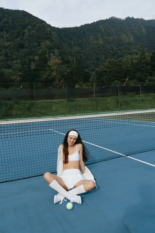 Young female tennis player in sporty outfit sitting on the court, expressing joy and relaxation against a lush mountain background