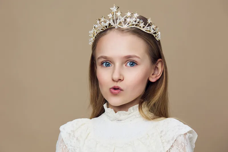 Young girl with a tiara making a playful expression against a beige background, dressed in a vintage white dress, perfect for children s portraits and celebrations