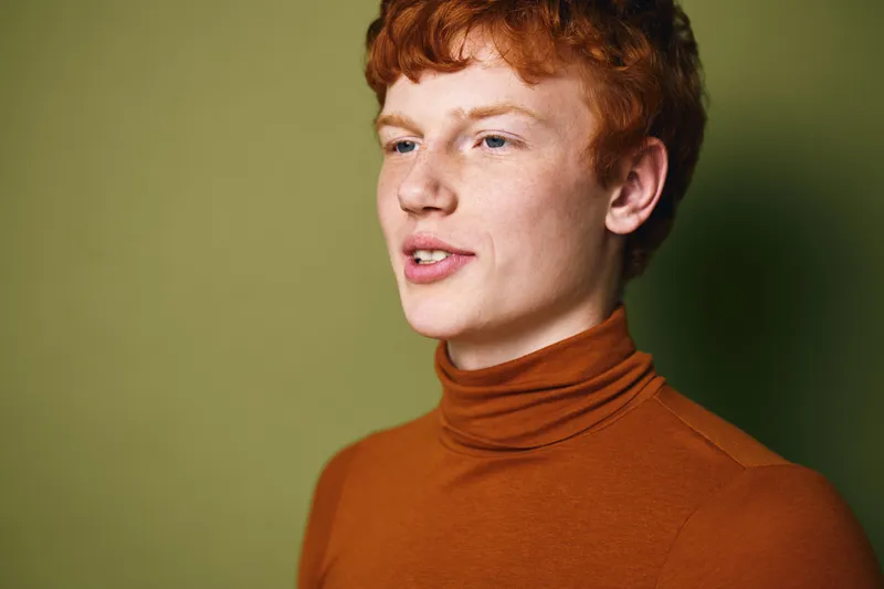 Young man with red hair wearing orange turtleneck sweater looking away with thoughtful expression on green background with soft lighting in studio portrait.