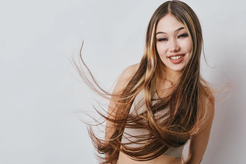 Smiling young woman with long flowing hair wearing casual top posing against white background with natural makeup and joyful expression in studio lighting.