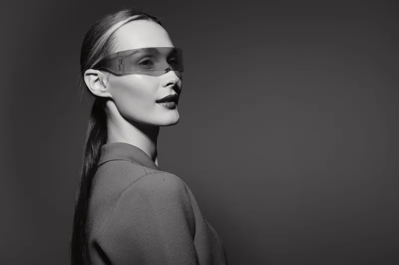 Portrait of a fair skinned European woman with slicked back hair wearing translucent visor glasses and textured jacket, looking confidently to the side on a dark studio background.