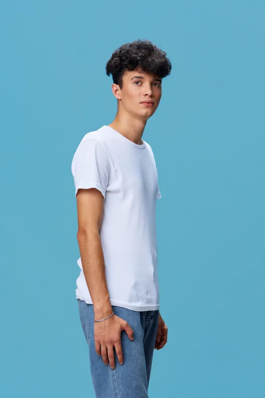 young man wearing a white t-shirt and blue jeans standing against a solid blue background, casual style, looking confident with curly hair
