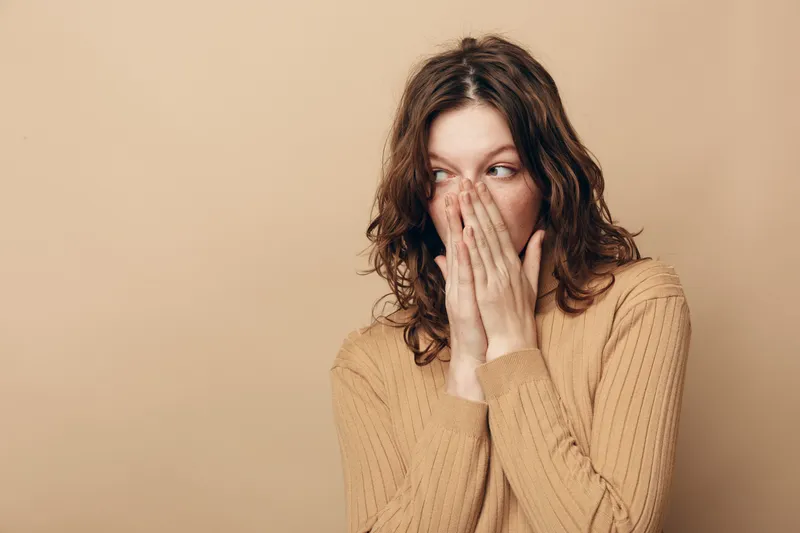 Young woman with brown hair wearing beige sweater covers her mouth with hands, looking sideways with surprised expression on beige background. Concept of shock, secrecy, and emotion.