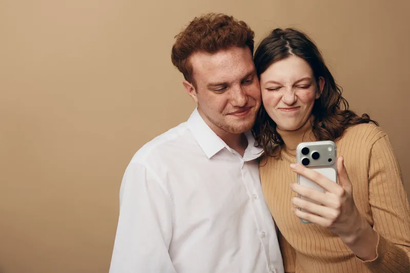 Young couple takes a selfie together, smiling and making funny faces. Casual clothing and warm beige background create a relaxed and playful mood. Concept of friendship and happiness.