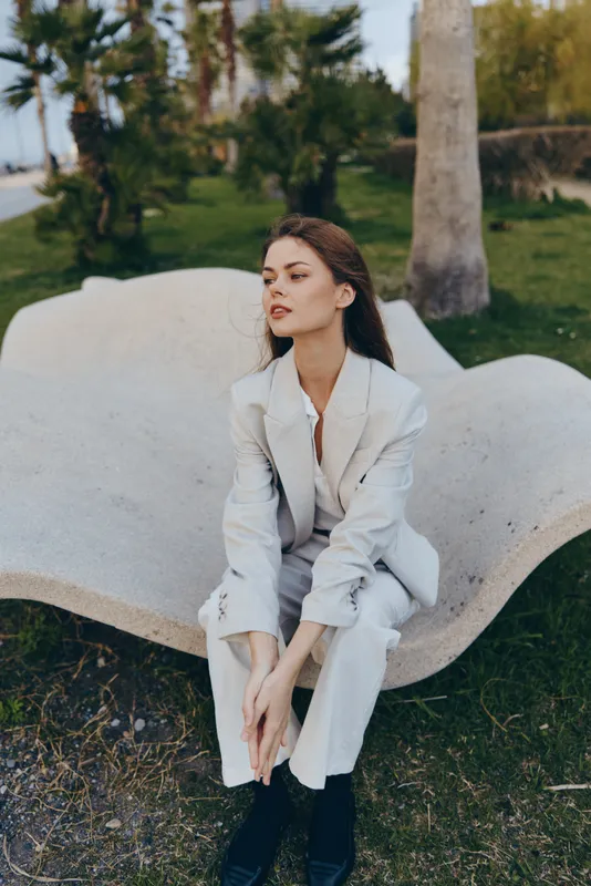 Woman in light gray pantsuit sitting outdoors on a modern bench, contemplative expression, natural green park background in spring sunlight