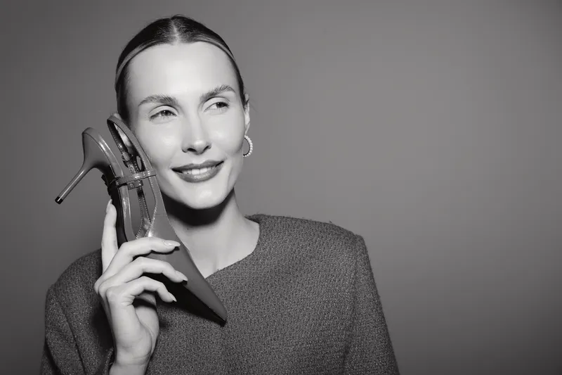 Smiling white woman with fair skin holds high heel shoe near face, looks away in studio. Concept for fashion, style, beauty, and elegance in monochrome portrait.