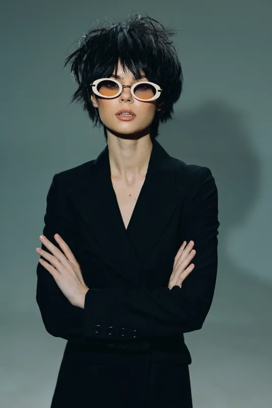 Stylish woman with short curly hair wearing white sunglasses and black blazer posing confidently against gray background