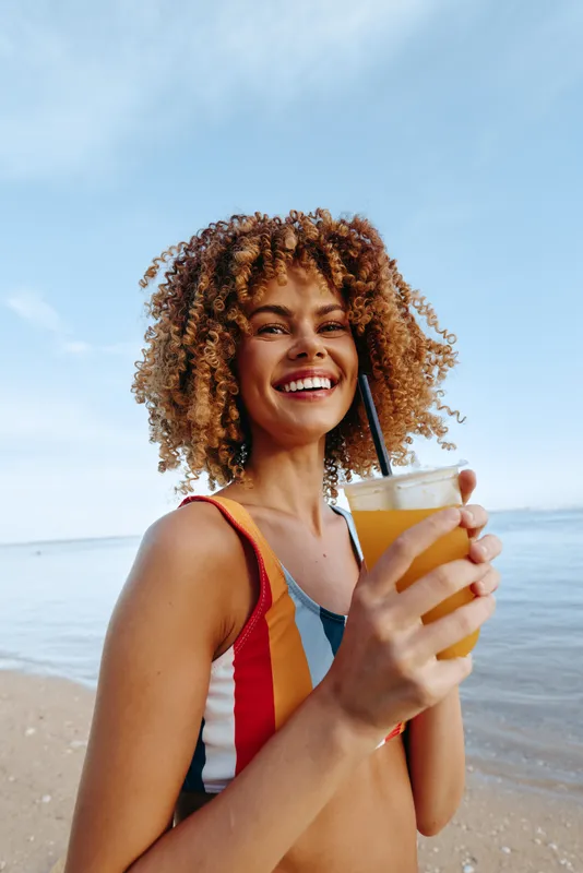 Smiling woman in a striped swimsuit holds a yellow drink on a sunny beach. Outdoor scene with sea and sky, conveying summer joy and carefree vibes.
