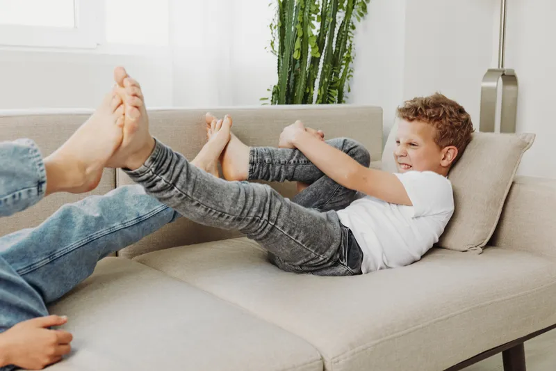 Playful young boy in white t-shirt and jeans laughs while holding feet of adult on beige sofa in bright living room. Concept of family fun and bonding.