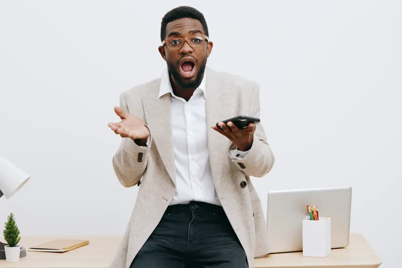 Excited young African American man wearing glasses and a beige blazer in a modern office, holding a smartphone with an expressive surprised face, standing near a desk with a laptop, white background, 