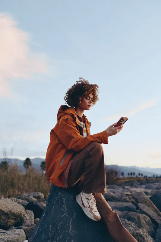 Woman in casual jacket with curly hair sitting on rocks outdoors, holding smartphone, mountains and sky in background, travel lifestyle of technology communication, natural daylight