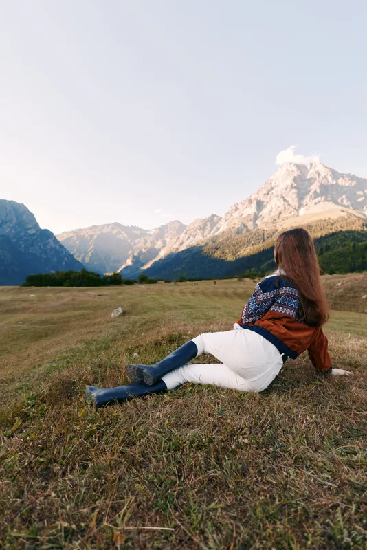 Woman in a meadow relaxing and watching mountains and dramatic landscape, nature escape with sweater and boots, outdoor travel scene in a peaceful alpine valley panorama.