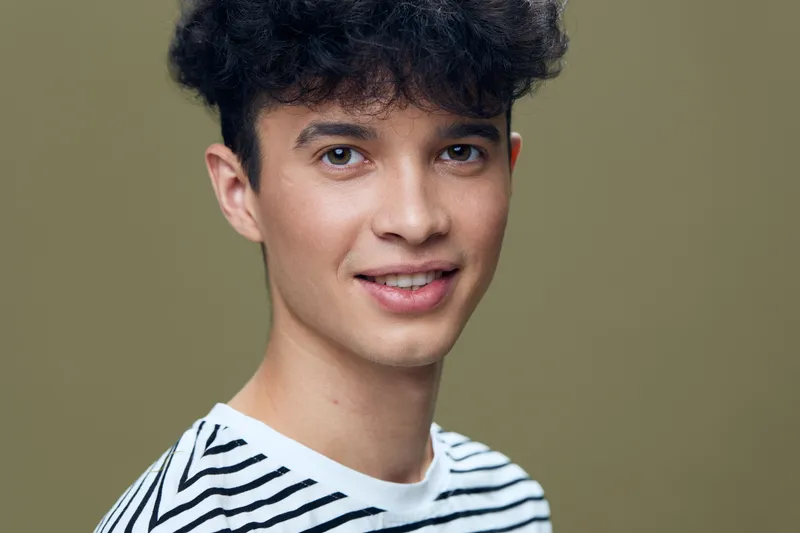 Smiling young man with curly hair wearing a black and white striped shirt against a simple beige background, expressing a relaxed and friendly mood.