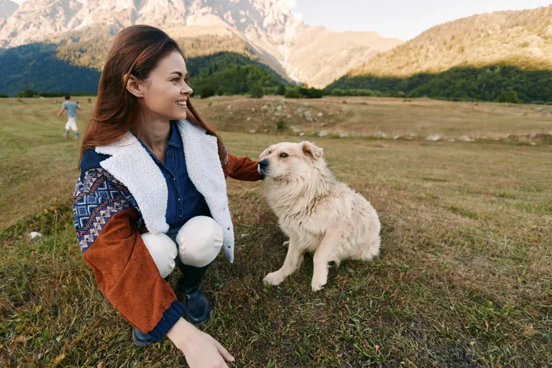 Woman smiling and petting a dog in a meadow by the mountains, outdoor nature portrait showing countryside companionship, joyful bonding and relaxed weekend adventure in grassy field.