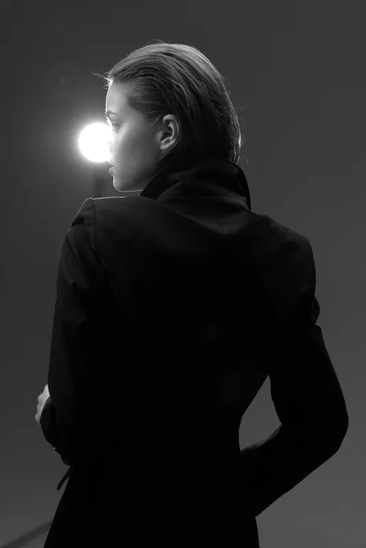 Black and white portrait of a woman in dark clothing with dramatic side lighting and moody atmosphere.