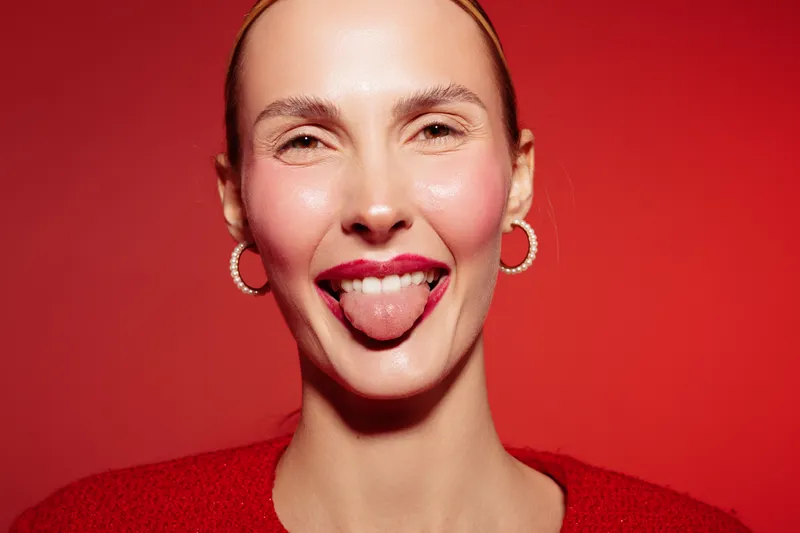 Close-up of a smiling woman with earrings sticking out her tongue against a red background. Concept of playful expression, fun, and confident female portrait in studio lighting.