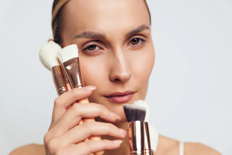 Young woman with natural makeup holds several makeup brushes close to her face while looking directly at the camera with a neutral expression.