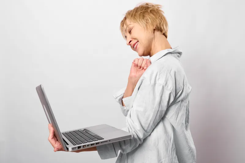 Woman laptop blonde smile technology remote work in office, side portrait on white background. She holds a computer and typing calmly, touching her neck, soft studio lighting.