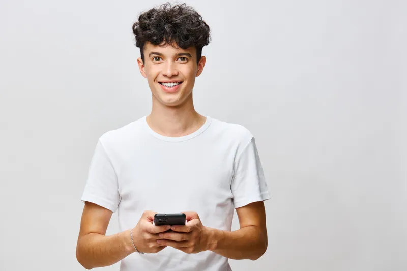 Улыбающийся молодой человек держит смартфон, стоя на фоне светло-серого фона, демонстрируя современные технологии и коммуникацию