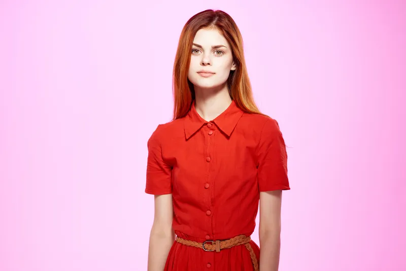 Рыжеволосая женщина в красном платье демонстрирует модный элегантный стиль на изолированном фоне