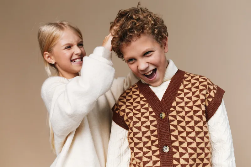 Two young friends pose in cozy knitwear, bright smiles and lively energy as one teases the other with a playful hair tug in a warm studio setting.