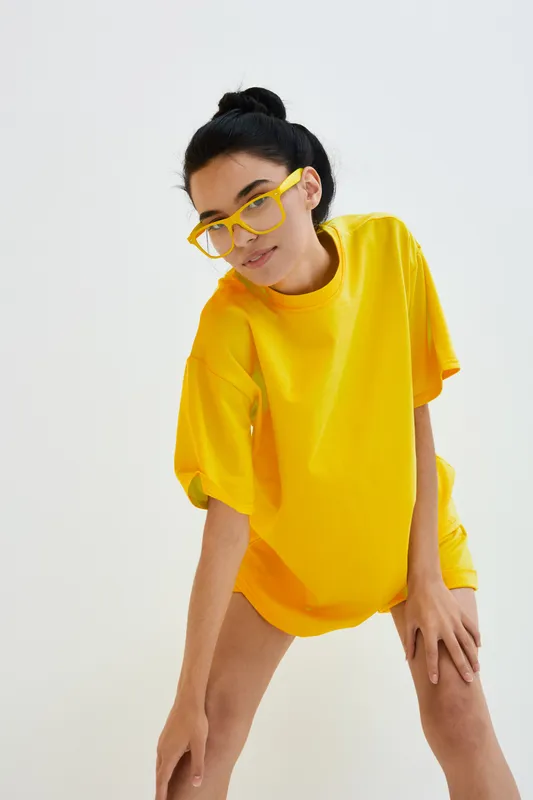 Young woman wearing oversized bright yellow shirt and matching glasses poses against white background, showcasing casual modern style with confident expression and black hair.