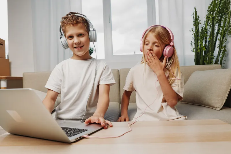 Happy boy and yawning girl wearing headphones using laptop at home. Cozy living room with natural light. Concept of children enjoying music and relaxation.