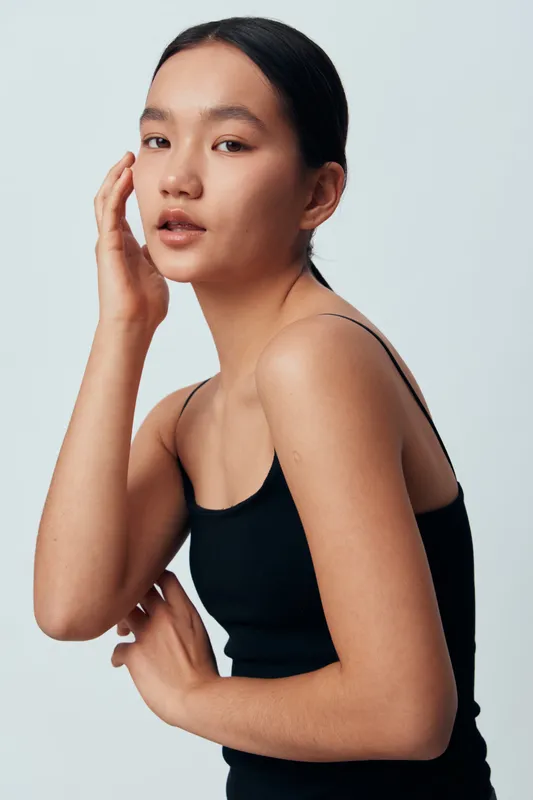 Young woman with flawless skin and minimal makeup posing in black tank top, natural beauty and confidence with hand on face against plain background in studio portrait.