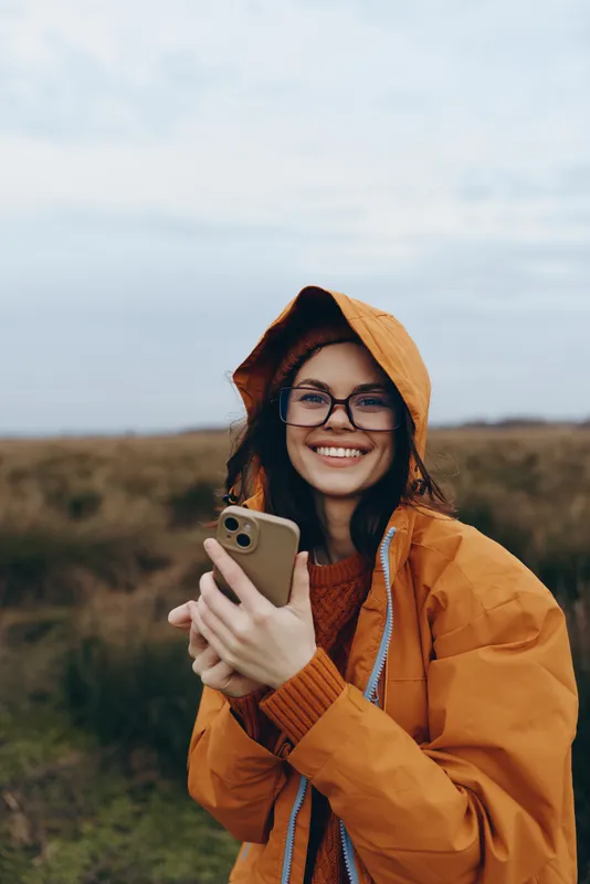 Happy woman lifestyle outdoors in autumn field, wearing orange hooded jacket and glasses, smiling while holding smartphone for selfie, natural light portrait, casual travel mood, soft background