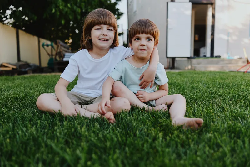 Two smiling boys sitting on green grass, enjoying a sunny day together, showing the joy of childhood and friendship in a cozy outdoor setting.