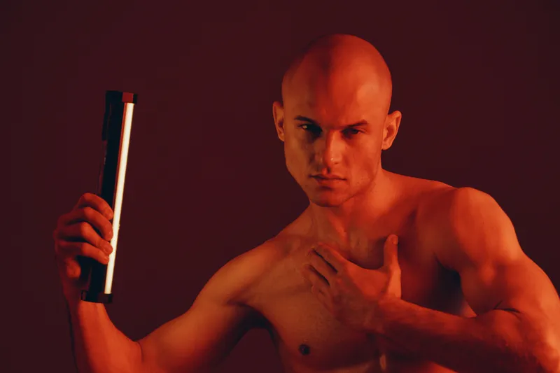 Strong man holding a flashlight, showcasing muscular physique against a dark red background. This captivating shot is striking and powerful, highlighting his intensity.