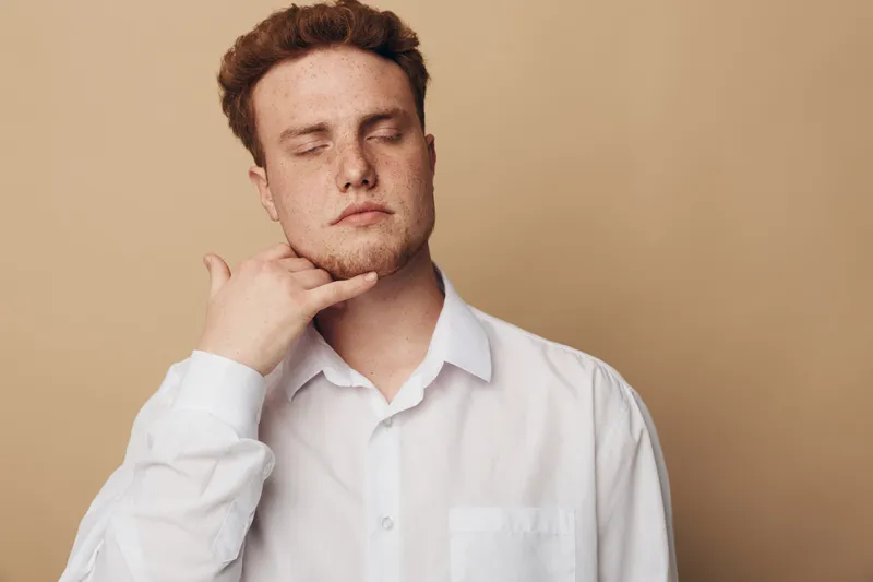 Young man in white shirt with closed eyes makes call me gesture with hand near face on beige background. Concept of communication, phone call, gesture, and casual style.